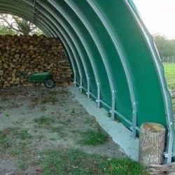 Vue intérieure d'une serre tunnel verte utilisée pour stocker du bois de chauffage avec une brouette à l'intérieur.