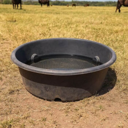 Bac de pâturage rond en plastique noir, rempli d’eau, posé sur une prairie sèche en été, avec plusieurs bovins visibles à l’arri
