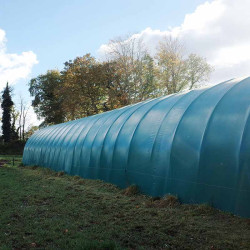 Vue latérale de l’abri tunnel Deschamps bâché en vert, installé en prairie.