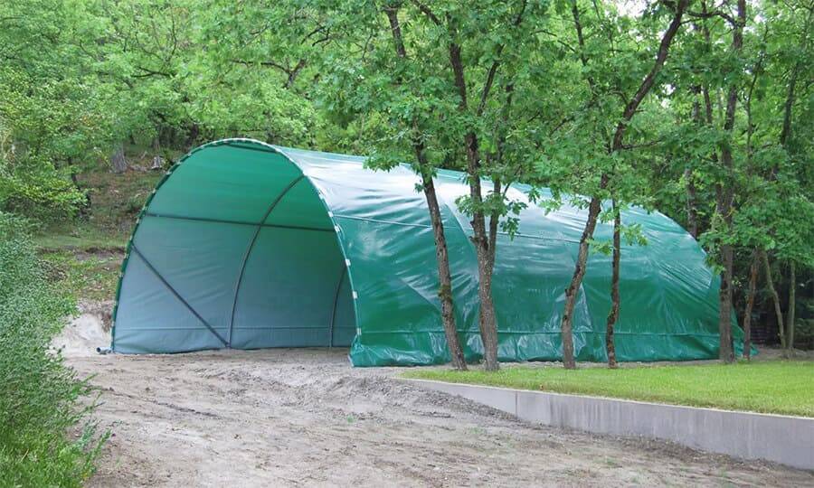 Un grand tunnel de stockage vert est érigé sur un terrain en terre, adossé à une forêt. Il est composé d'une structure métallique recouverte d'une bâche verte et est ouvert à une extrémité.