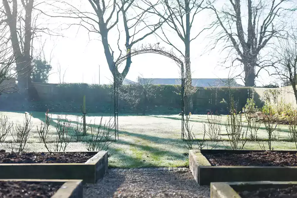 Vue d'un grand jardin anglais en hiver, avec des parterres surélevés, des arbres sans feuilles, et une arche en métal, sous une lumière matinale douce.