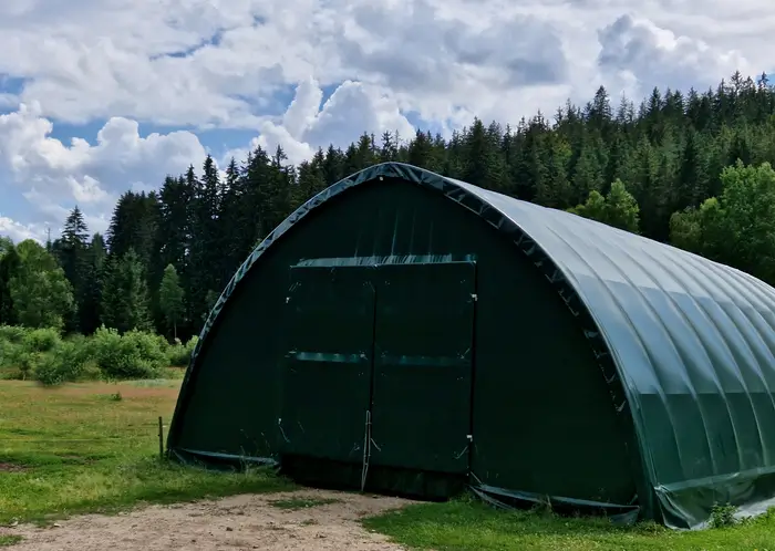 Un grand abri de stockage vert installé dans une clairière herbeuse, entouré d'une forêt dense sous un ciel partiellement nuageux.