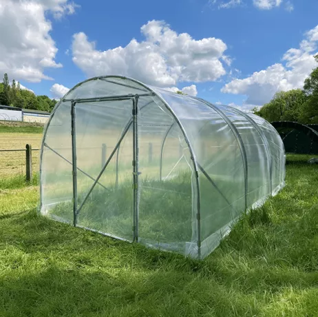 Une serre tunnel avec une structure en plastique transparent et des arceaux métalliques, installée sur un terrain herbeux sous un ciel bleu parsemé de nuages.