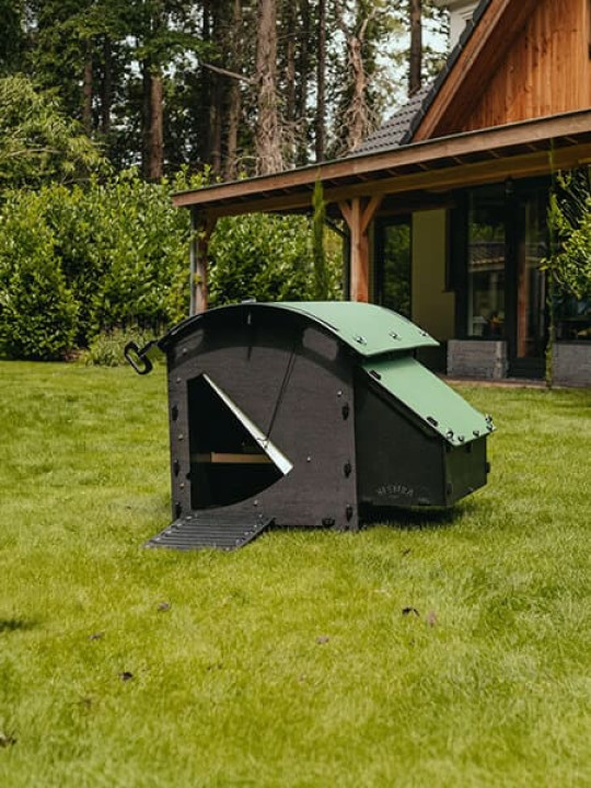 Poulailler moderne en plastique recyclé dans un jardin, avec toit vert incliné et rampe d'accès, placé sur une pelouse verte devant une maison en bois entourée de végétation.